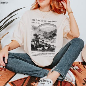 Puede incluir: Una mujer lleva una camiseta de color crema con el texto "The Lord is my shepherd: Psalm 23:1-6" y una ilustraci&oacute;n en blanco y negro de un pastor y ovejas. Lleva pantalones vaqueros azules y un collar de oro.
