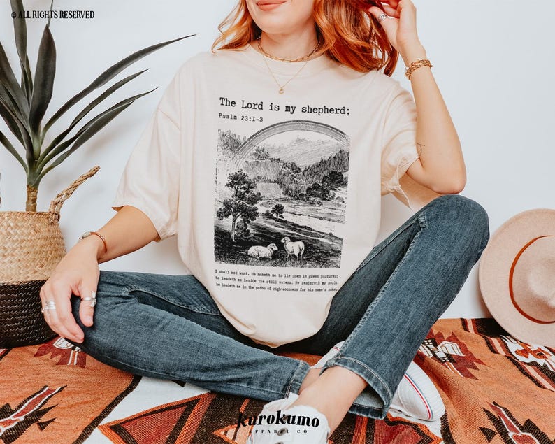 Puede incluir: Una mujer lleva una camiseta de color crema con el texto "The Lord is my shepherd: Psalm 23:1-6" y una ilustraci&oacute;n en blanco y negro de un pastor y ovejas. Lleva pantalones vaqueros azules y un collar de oro.