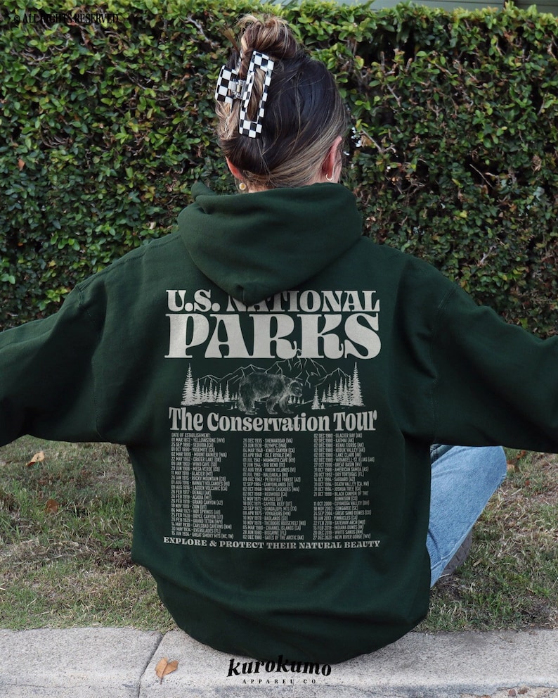 Pu&ograve; includere: Felpa con cappuccio verde scuro con la scritta "U.S. NATIONAL PARKS" e "The Conservation Tour" sul retro. Il design include un orso e un elenco di date dei parchi. La felpa con cappuccio &egrave; indossata da una persona.