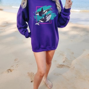 Puede incluir: Sudadera morada con un estampado gr&aacute;fico de dos orcas blancas y negras nadando en agua turquesa con pinceladas rosas y blancas. La sudadera la lleva puesta una mujer de pie en una playa de arena.