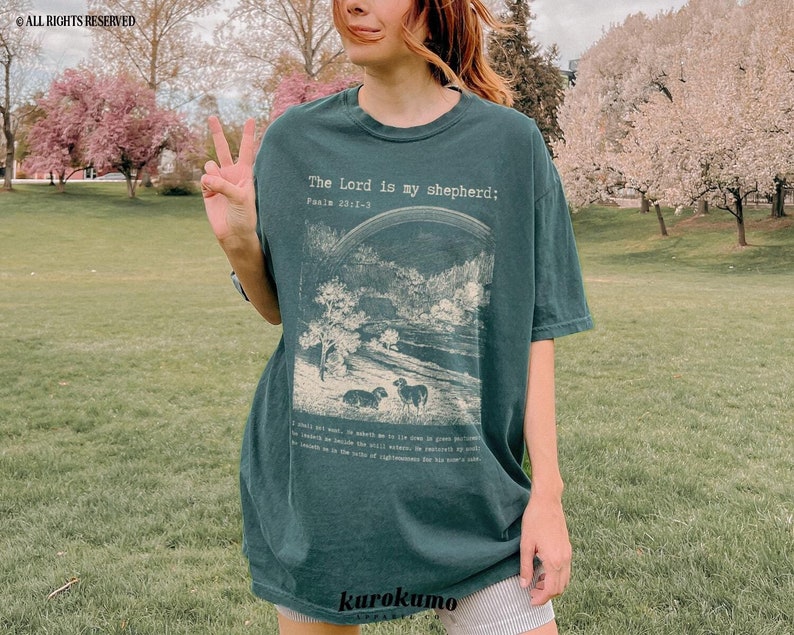 Puede incluir: Una camiseta verde oscuro con una ilustraci&oacute;n vintage de un pastor y ovejas. El texto "The Lord is my shepherd; Psalm 23:1-3" est&aacute; impreso en la parte delantera de la camiseta.