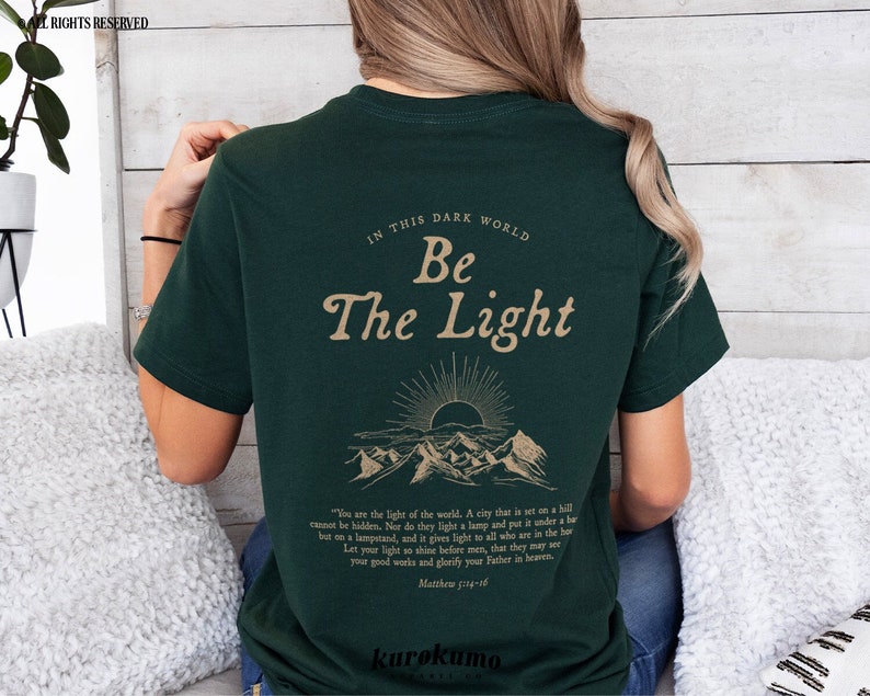 May include: A dark green t-shirt with a gold graphic of mountains and a sunrise. The text "In this dark world, Be the Light" is printed on the shirt. The text "You are the light of the world. A city that is set on a hill cannot be hidden. Nor do they light a lamp and put it under a bushel, but on a lampstand, and it gives light to all who are in the house. Let your light so shine before men, that they may see your good works and glorify your Father in heaven. Matthew 5:14-16" is printed below the graphic.