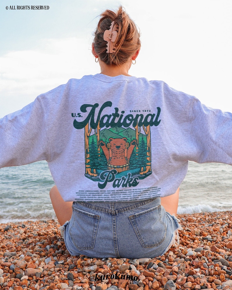 Może przedstawiać: Jasnoszara bluza z grafiką niedźwiedzia w leśnej scenerii, z napisem "U.S. National Parks Since 1972" w kolorze zielonym i białym. Bluza jest noszona przez osobę siedzącą na kamienistej plaży.