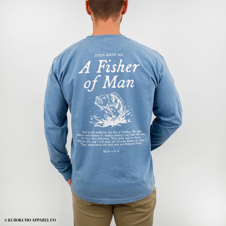 May include: A light blue long-sleeved shirt featuring the text "JESUS MADE ME A Fisher of Man" in white. A white graphic of a fish leaping from water is below the text. The shirt also has a Bible verse printed on it.