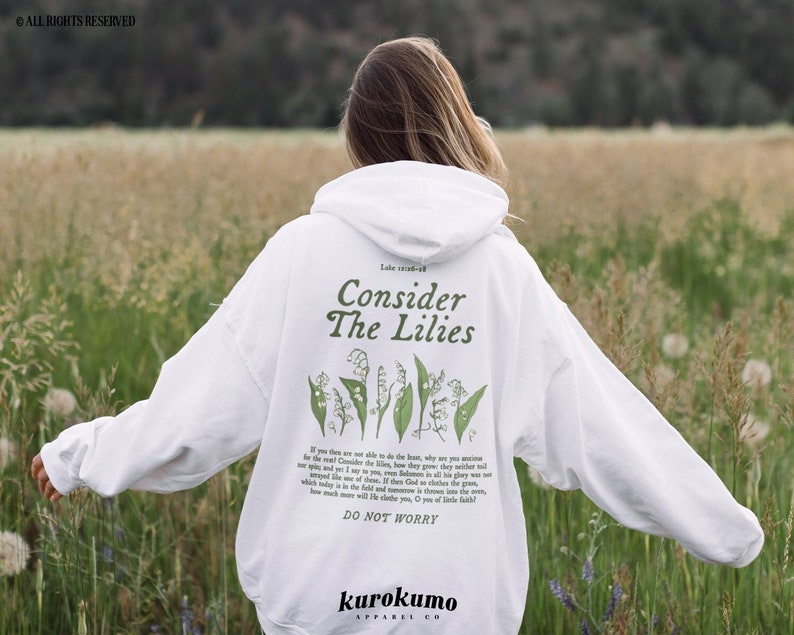 Pu&ograve; includere: Felpa con cappuccio bianca con il testo "Consider The Lilies" e una citazione dalla Bibbia, Luca 12:27, stampata sul retro. Il testo &egrave; in nero e la citazione &egrave; in grigio. La felpa &egrave; indossata da una persona in piedi in un campo di erba verde.