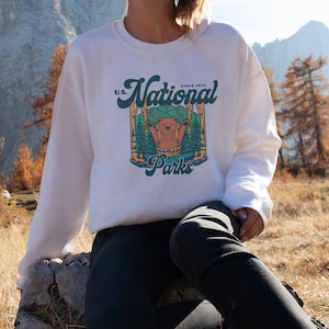 Może przedstawiać: Biała bluza z okrągłym dekoltem z grafiką przedstawiającą niedźwiedzia w leśnej scenerii z napisem "U.S. National Parks Since 1872". Projekt jest w odcieniach zieleni, brązu i pomarańczy. Bluza jest w stylu casual.