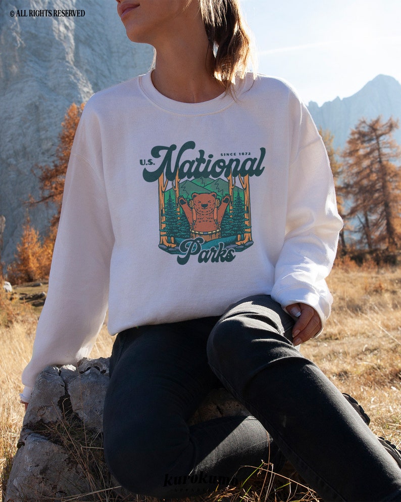 Może przedstawiać: Biała bluza z okrągłym dekoltem z grafiką przedstawiającą niedźwiedzia w leśnej scenerii z napisem "U.S. National Parks Since 1872". Projekt jest w odcieniach zieleni, brązu i pomarańczy. Bluza jest w stylu casual.