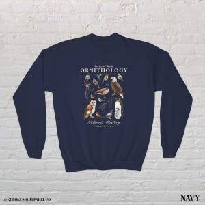 May include: Navy blue sweatshirt featuring a detailed illustration of various bird species. The design includes the text "Study of Birds ORNITHOLOGY Natural History Collection". The sweatshirt is a comfortable, long-sleeved garment.