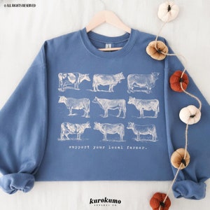May include: A blue sweatshirt with a white graphic of nine cows and the text "support your local farmer." The sweatshirt is hanging on a wooden hanger against a white background.