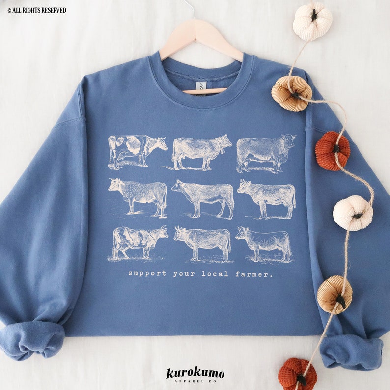 May include: A blue sweatshirt with a white graphic of nine cows and the text "support your local farmer." The sweatshirt is hanging on a wooden hanger against a white background.