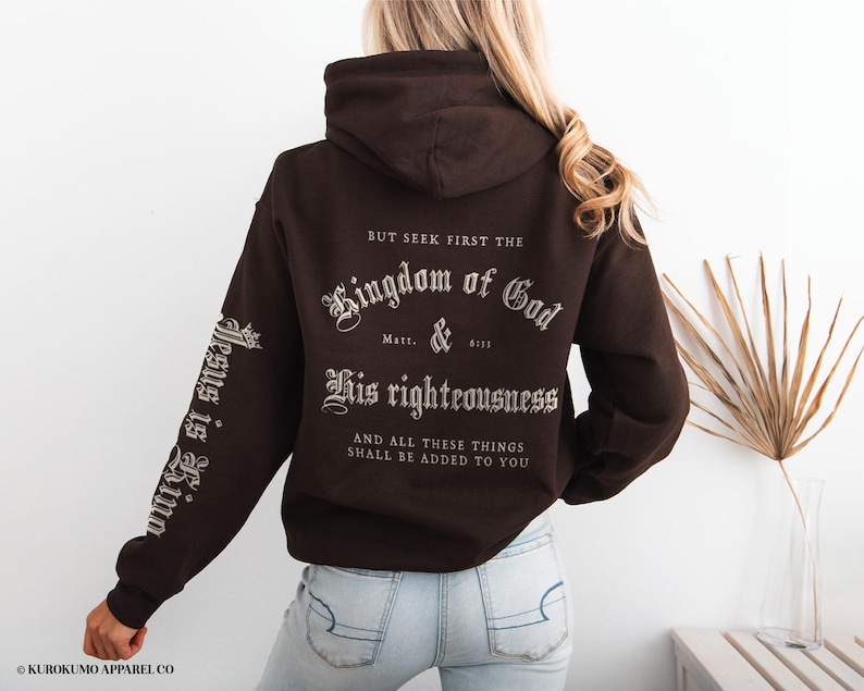 Op de afbeelding: Een bruine hoodie met een witte grafische print die "BUT SEEK FIRST THE Kingdom of God Matt. 6:33 His righteousness AND ALL THESE THINGS SHALL BE ADDED TO YOU" en de tekst "Jesus is King" op de mouw staat.