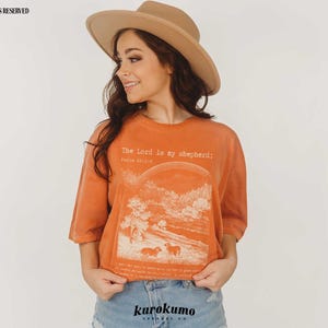 Puede incluir: Una mujer con una camiseta naranja quemada con el texto "The Lord is my shepherd; Psalm 23:1-6" y un gr&aacute;fico de un paisaje con ovejas. Lleva un sombrero marr&oacute;n y unos vaqueros azules.