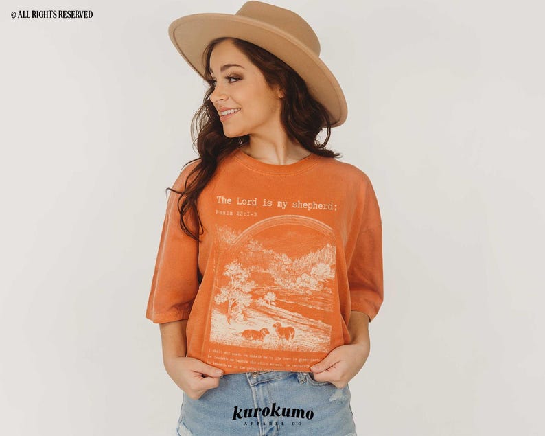 Puede incluir: Una mujer con una camiseta naranja quemada con el texto "The Lord is my shepherd; Psalm 23:1-6" y un gr&aacute;fico de un paisaje con ovejas. Lleva un sombrero marr&oacute;n y unos vaqueros azules.