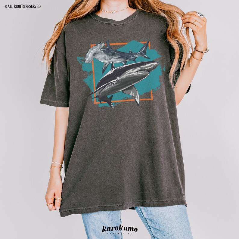 May include: A dark gray t-shirt with a graphic print of two sharks, a hammerhead and a great white, on a teal blue background with orange accents. The graphic is framed in a square.
