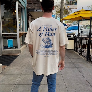 May include: A cream-coloured t-shirt with a blue graphic of a fish jumping out of the water. The text "Jesus Made Me A Fisher of Man" is printed on the shirt. The text "And as He walked by the Sea of Galilee, He saw Simon and Andrew his brother casting a net into the sea.  They immediately left their nets and followed Him." is printed below the graphic.  The shirt is being worn by a person.