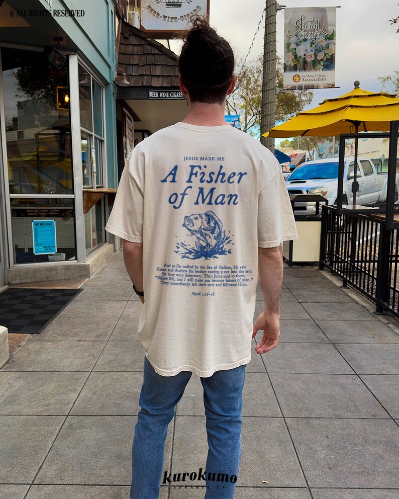 May include: A cream-coloured t-shirt with a blue graphic of a fish jumping out of the water. The text "Jesus Made Me A Fisher of Man" is printed on the shirt. The text "And as He walked by the Sea of Galilee, He saw Simon and Andrew his brother casting a net into the sea.  They immediately left their nets and followed Him." is printed below the graphic.  The shirt is being worn by a person.