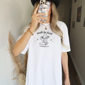 Pu&ograve; includere: Maglietta bianca con un'illustrazione in bianco e nero di uno stivale da cowboy con il testo "walk by faith" e "2 Corinthians 5:7".