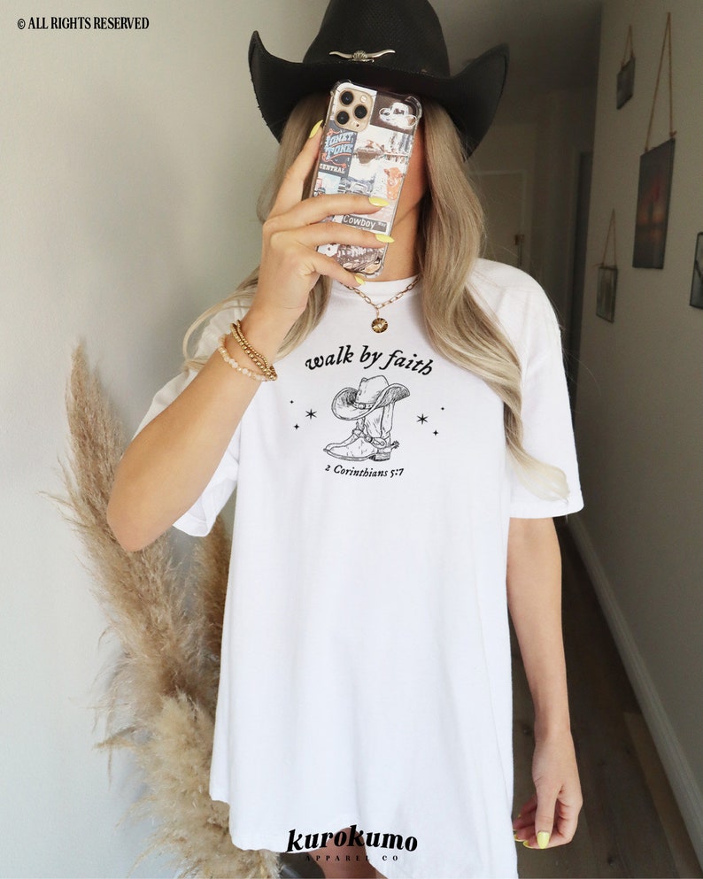 Pu&ograve; includere: Maglietta bianca con un'illustrazione in bianco e nero di uno stivale da cowboy con il testo "walk by faith" e "2 Corinthians 5:7".