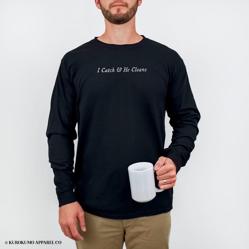 May include: Black long-sleeved shirt with the white text "I Catch & He Cleans". The shirt is worn with khaki trousers. A white mug is held in the hand. The brand name "KUROKUMO APPAREL CO" is at the bottom.
