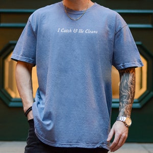 May include: A blue t-shirt with the text "I Catch & He Cleans" printed in white on the front. The shirt has a relaxed fit and a slightly longer length.