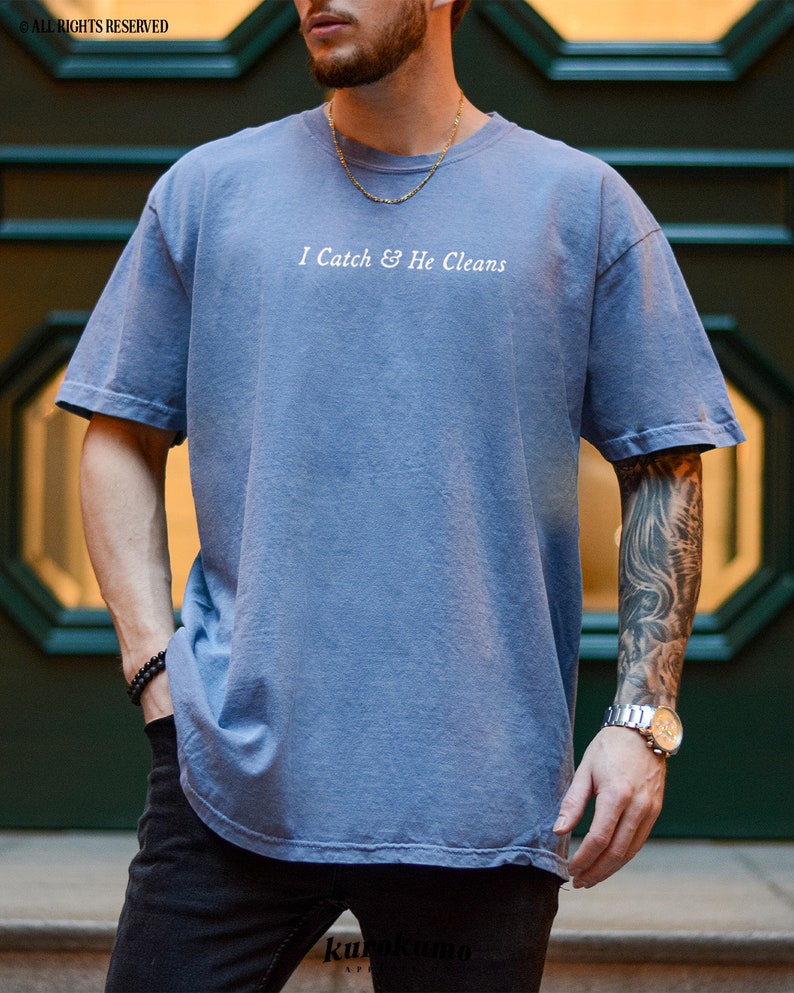 May include: A blue t-shirt with the text "I Catch & He Cleans" printed in white on the front. The shirt has a relaxed fit and a slightly longer length.