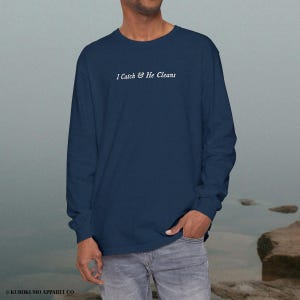 May include: A navy blue long-sleeved shirt featuring the white text "I Catch & He Cleans". The shirt is made of a soft fabric and is suitable for casual wear. The brand name "KUROKUMO APPAREL CO." is printed at the bottom.