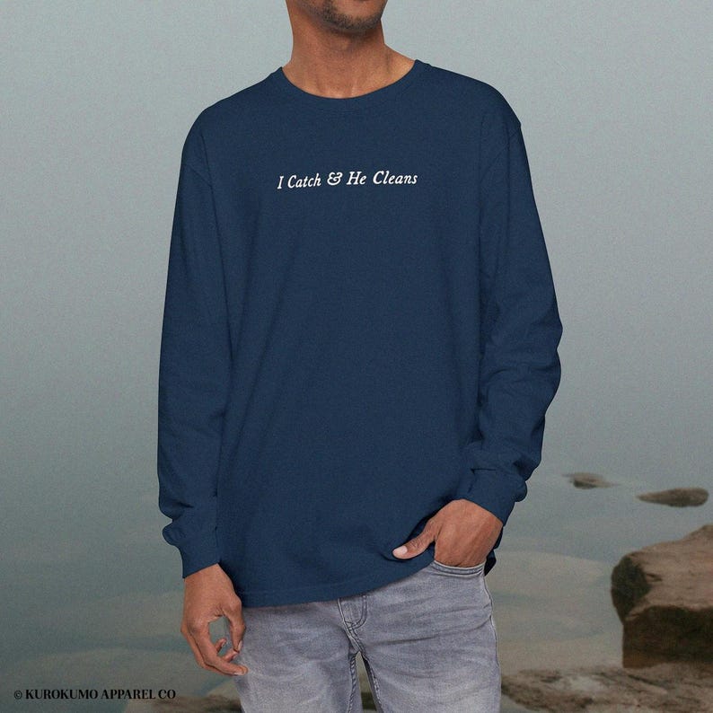 May include: A navy blue long-sleeved shirt featuring the white text "I Catch & He Cleans". The shirt is made of a soft fabric and is suitable for casual wear. The brand name "KUROKUMO APPAREL CO." is printed at the bottom.