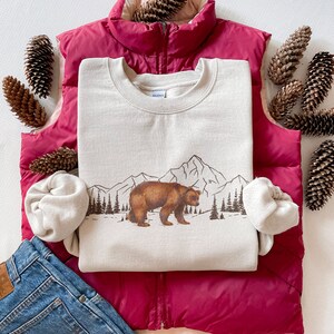 May include: A beige sweatshirt featuring a brown bear graphic walking in front of a mountain range and pine trees. The sweatshirt is layered on a magenta vest, with blue jeans and pine cones. The text "ALL RIGHTS RESERVED" is visible.