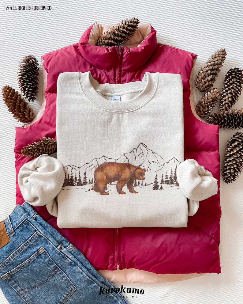 May include: A beige sweatshirt featuring a brown bear graphic walking in front of a mountain range and pine trees. The sweatshirt is layered on a magenta vest, with blue jeans and pine cones. The text "ALL RIGHTS RESERVED" is visible.