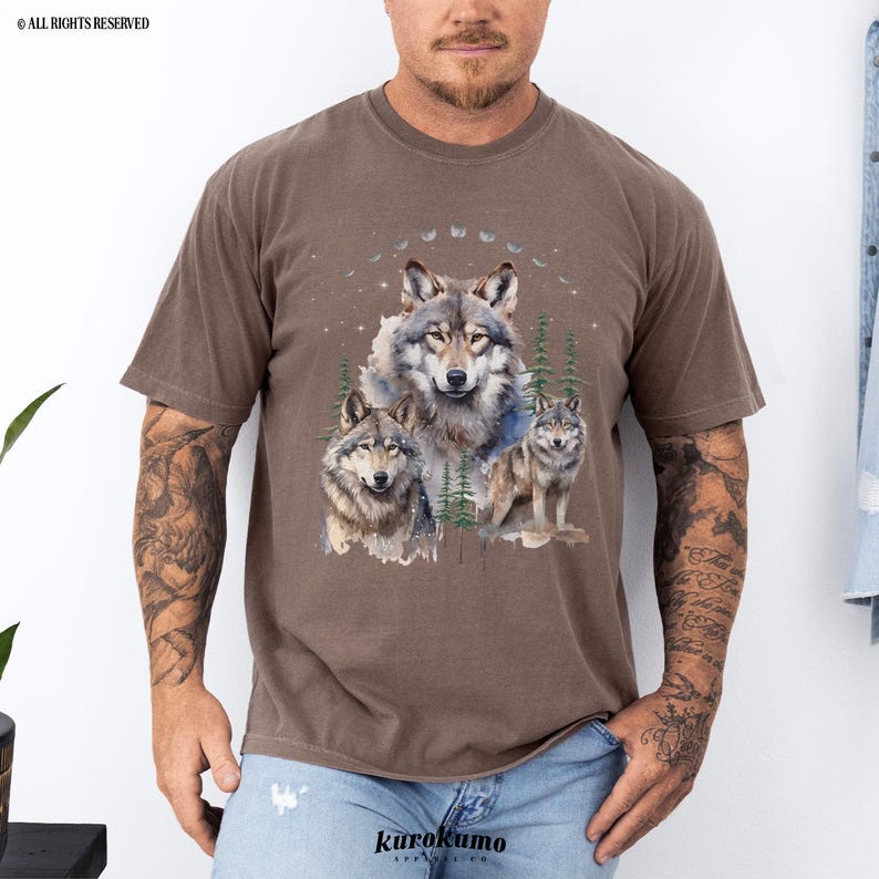 May include: A brown t-shirt with a watercolor design of three wolves in a forest setting. The wolves are gray and white with a blue and green background.