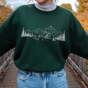 May include: A dark green sweatshirt featuring a white graphic of a bear, mountains, and trees. The sweatshirt has a crew neck and long sleeves. The brand name "kurokumo" is printed at the bottom. The words "ALL RIGHTS RESERVED" are printed at the top.
