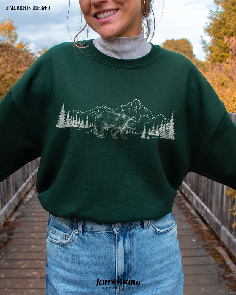 May include: A dark green sweatshirt featuring a white graphic of a bear, mountains, and trees. The sweatshirt has a crew neck and long sleeves. The brand name "kurokumo" is printed at the bottom. The words "ALL RIGHTS RESERVED" are printed at the top.