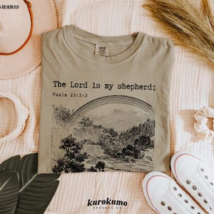Puede incluir: Una camiseta verde claro con el texto "The Lord is my shepherd; Psalm 23:1-3" impreso en la parte delantera. La camiseta presenta una ilustraci&oacute;n en blanco y negro de un arco&iacute;ris y un paisaje.