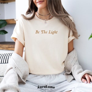 May include: A white t-shirt with the text "Be The Light" in brown lettering. The shirt is being worn by a person with long brown hair.