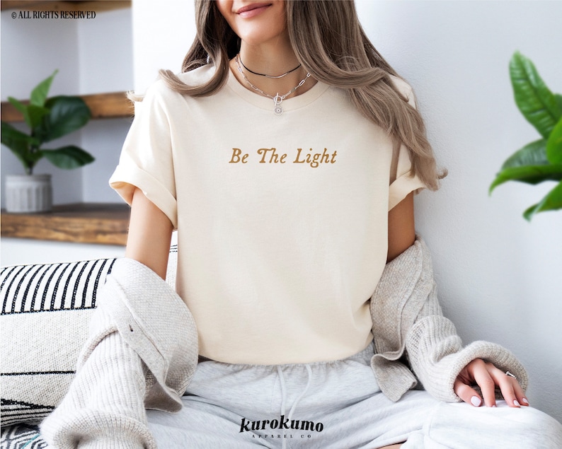 May include: A white t-shirt with the text "Be The Light" in brown lettering. The shirt is being worn by a person with long brown hair.
