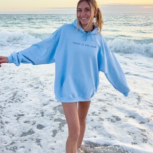 May include: A light blue hoodie with the text "Trust in the Lord" printed on the front. The hoodie is worn by a person standing on a beach with white sand and blue water.