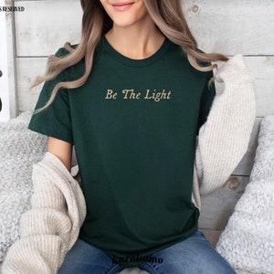 May include: A dark green t-shirt with the text "Be The Light" in a gold script font.