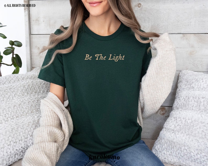 May include: A dark green t-shirt with the text "Be The Light" in a gold script font.