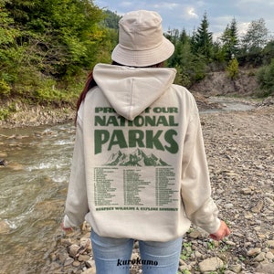 May include: Beige hoodie with the text "PROTECT OUR NATIONAL PARKS" in green, along with a list of national parks and mountain graphic. The hoodie is worn with a matching beige bucket hat and blue jeans. The image is taken outdoors.