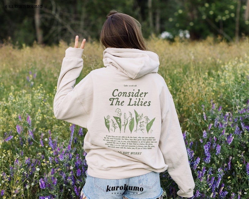Pu&ograve; includere: Una felpa con cappuccio beige con il testo "Consider the Lilies" e un'illustrazione floreale sul retro. La felpa &egrave; indossata da una persona in un campo di fiori viola.