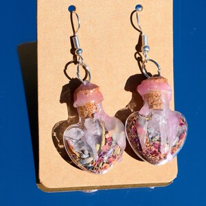 May include: A pair of heart-shaped glass bottle earrings with cork stoppers. The bottles are filled with pink and white crystals, dried flowers, and herbs. The earrings are hanging from silver hooks.