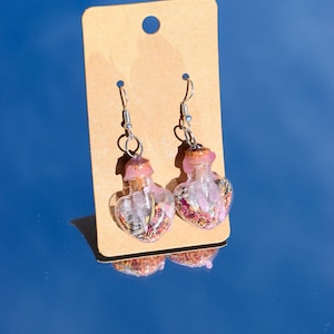 May include: A pair of silver heart-shaped earrings with clear glass bottles filled with pink and white crystals and dried flowers.