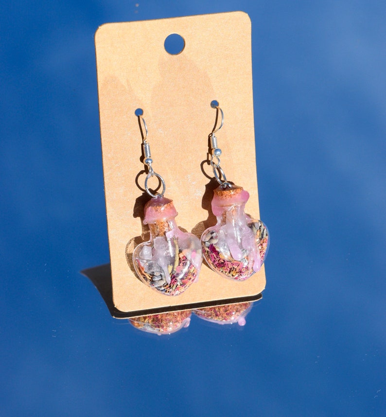 May include: A pair of silver heart-shaped earrings with clear glass bottles filled with pink and white crystals and dried flowers.