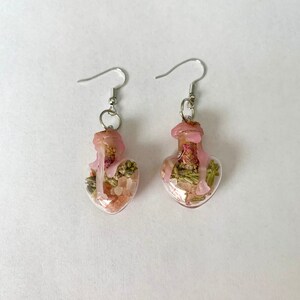 May include: A pair of heart-shaped glass bottle earrings with pink and green dried flowers and pink resin inside. The earrings have silver hooks.