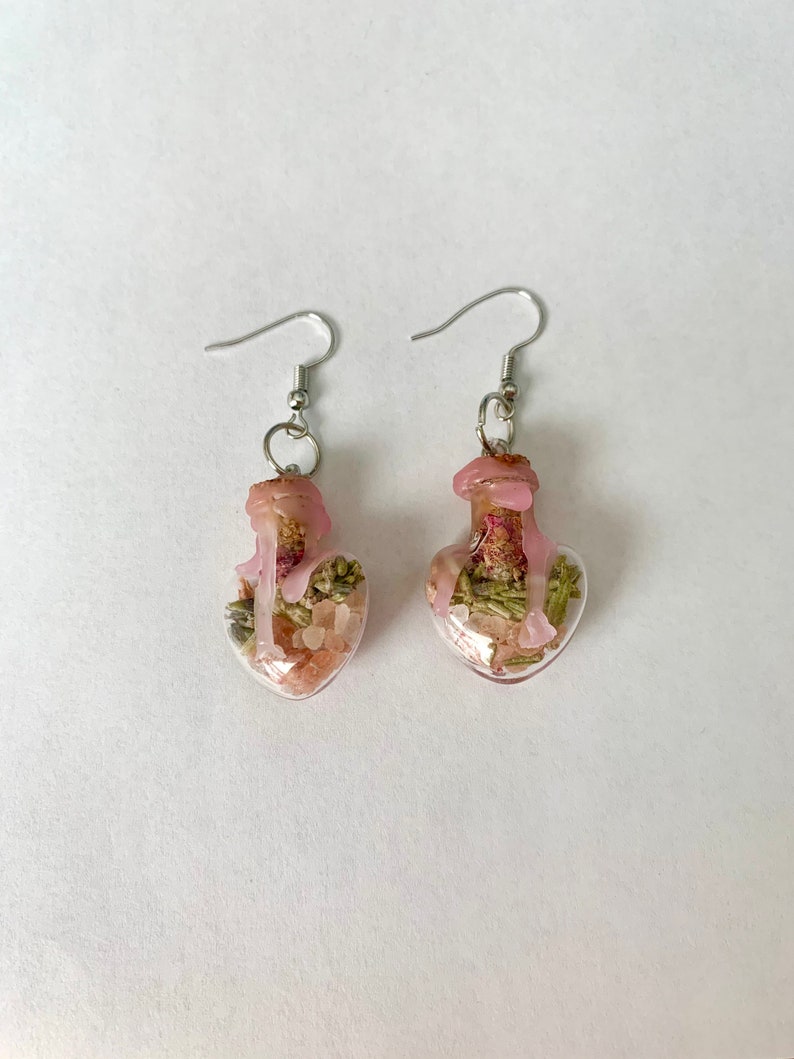 May include: A pair of heart-shaped glass bottle earrings with pink and green dried flowers and pink resin inside. The earrings have silver hooks.