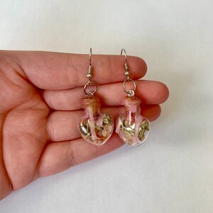 May include: A pair of heart-shaped glass bottle earrings with cork stoppers. The bottles are filled with dried herbs and pink crystals.