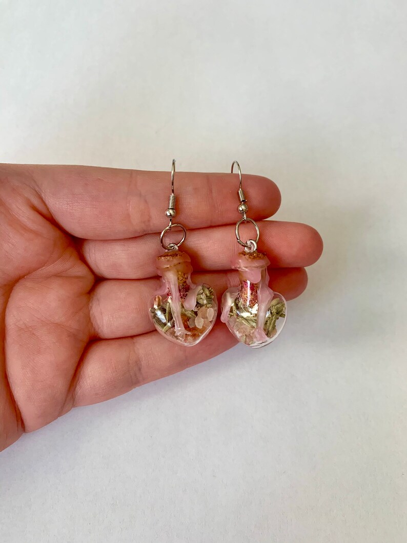 May include: A pair of heart-shaped glass bottle earrings with cork stoppers. The bottles are filled with dried herbs and pink crystals.