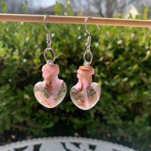 May include: A pair of heart-shaped glass bottle earrings with pink wax and dried flowers inside. The earrings are silver and have a hook closure.