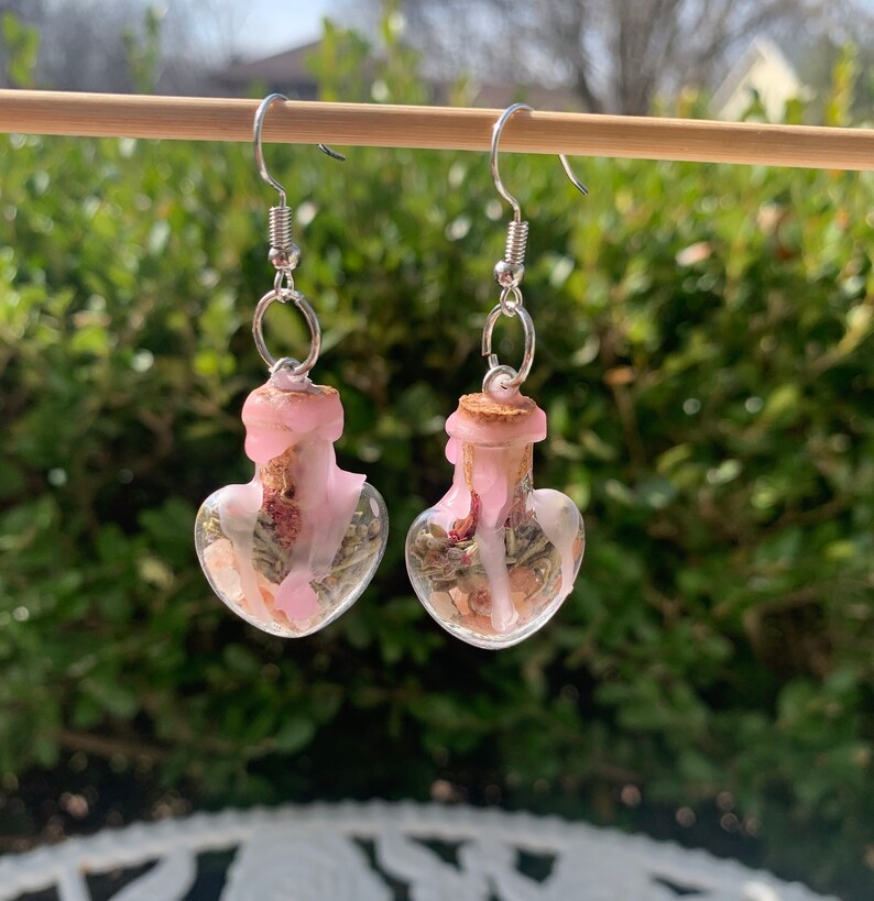 May include: A pair of heart-shaped glass bottle earrings with pink wax and dried flowers inside. The earrings are silver and have a hook closure.
