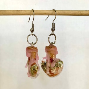 May include: A pair of pink resin earrings with clear glass bottles filled with dried herbs and crystals. The bottles are shaped like hearts and are attached to silver hooks.